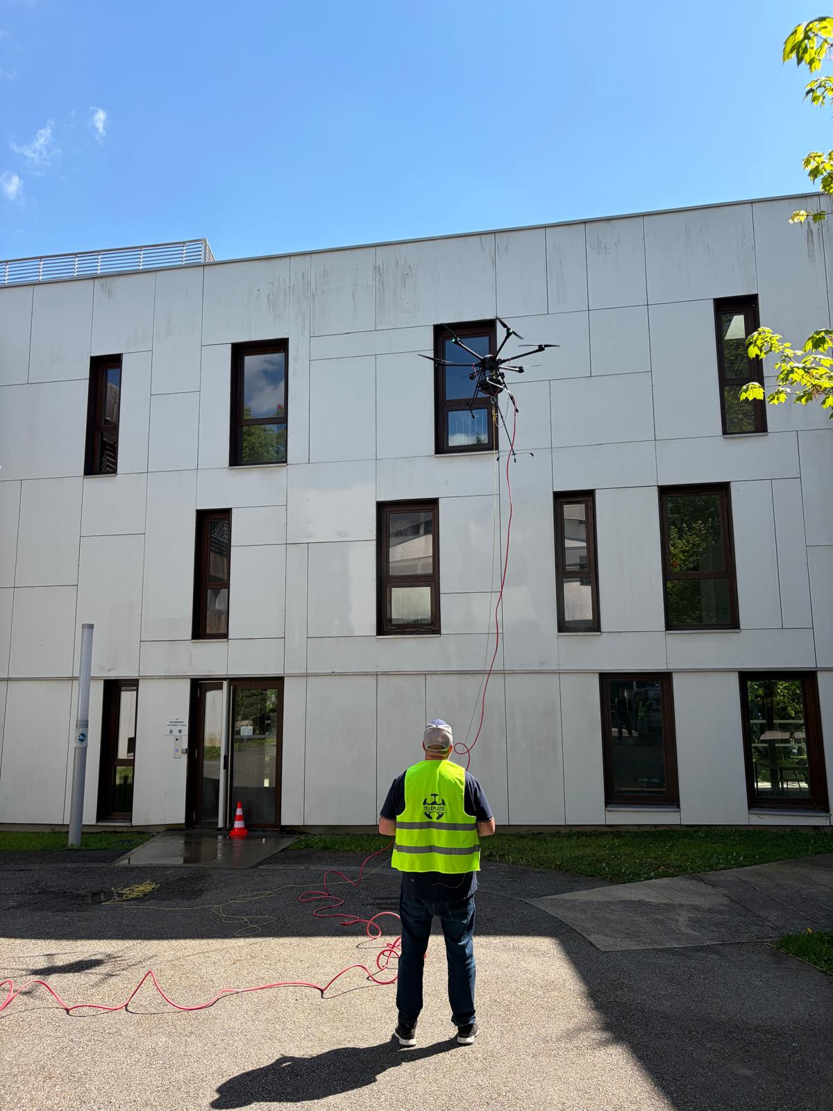 Technician in a neon safety vest watches a large multi-rotor drone hung by cables near a modern gray building window.