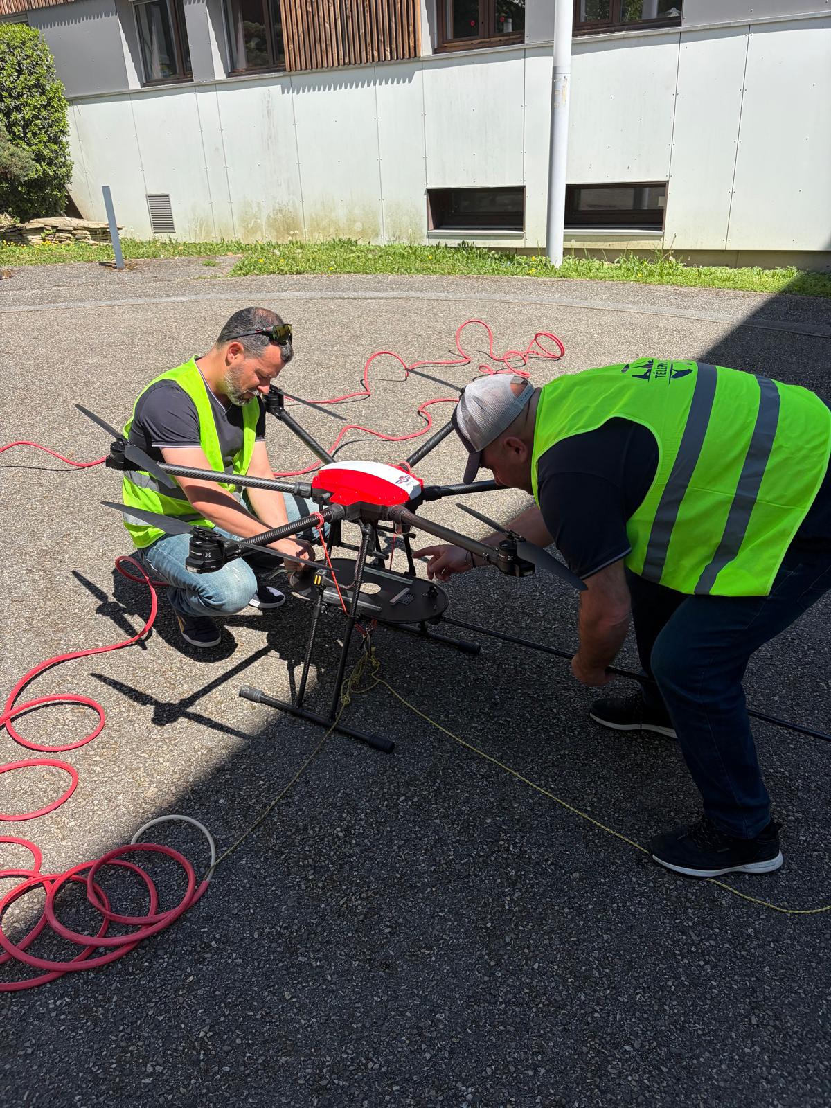 Two workers in neon safety vests crouch beside a red quadcopter drone on a paved area, adjusting controls with cables laid out nearby.”] ,